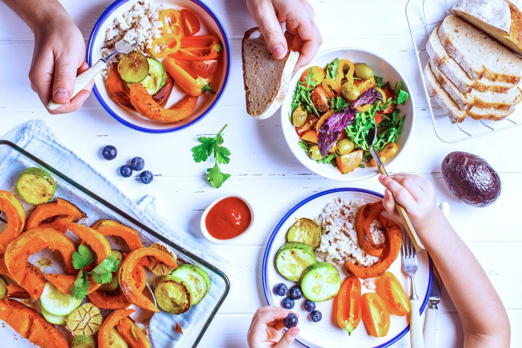 Ein Foto eines Tisches von oben mit gegrilltem Gemüse, Brot und Salat darauf und den Händen von zwei Menschen, die sich gegenübersitzen und essen.