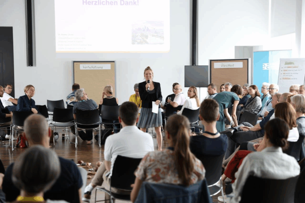 Aufnahme aus einer von KLUG organisierten Veranstaltung. Menschen sitzen in einem Raum im Kreis und in der Mitte steht die Referierende von KLUG und hält die Veranstaltung.