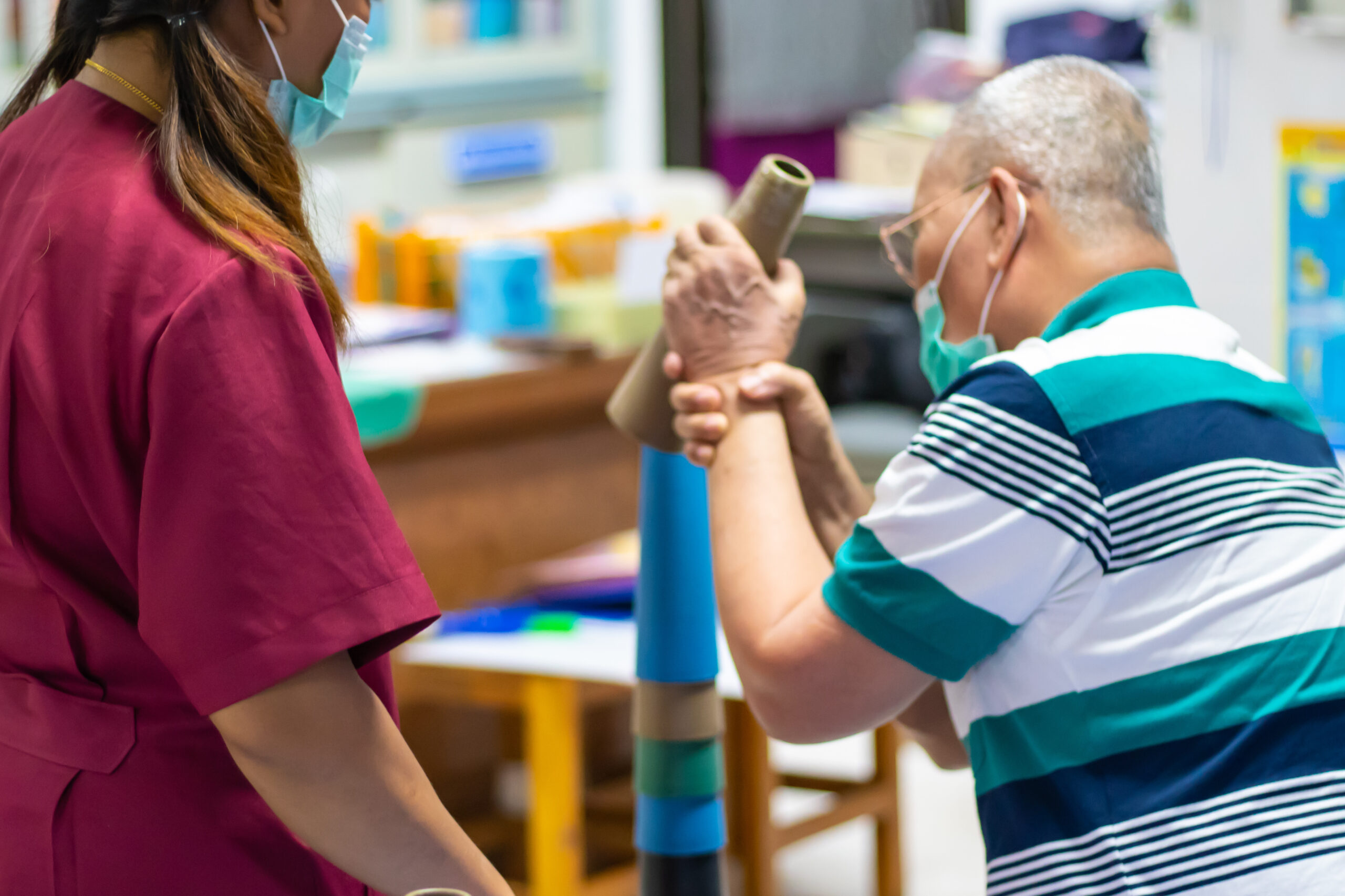 Ergotherapeuten gibt Patienten in gestreiftem Oberteil Handfunktionstraining unter Verwendung von Stapelkegeln in einem Therapieraum.
